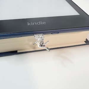 May include: A silver paperclip shaped like a bird is holding the pages of a book closed. The book is closed and the cover is cream coloured. The book is sitting on a blue surface. The book has the word "kindle" printed on the cover.