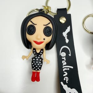 May include: A Coraline-themed keyring featuring a figurine with button eyes, a black dress with white polka dots, and red boots. The keyring includes a black strap with the name "Coraline" and a bat graphic. The keyring is attached to a gold-coloured ring.