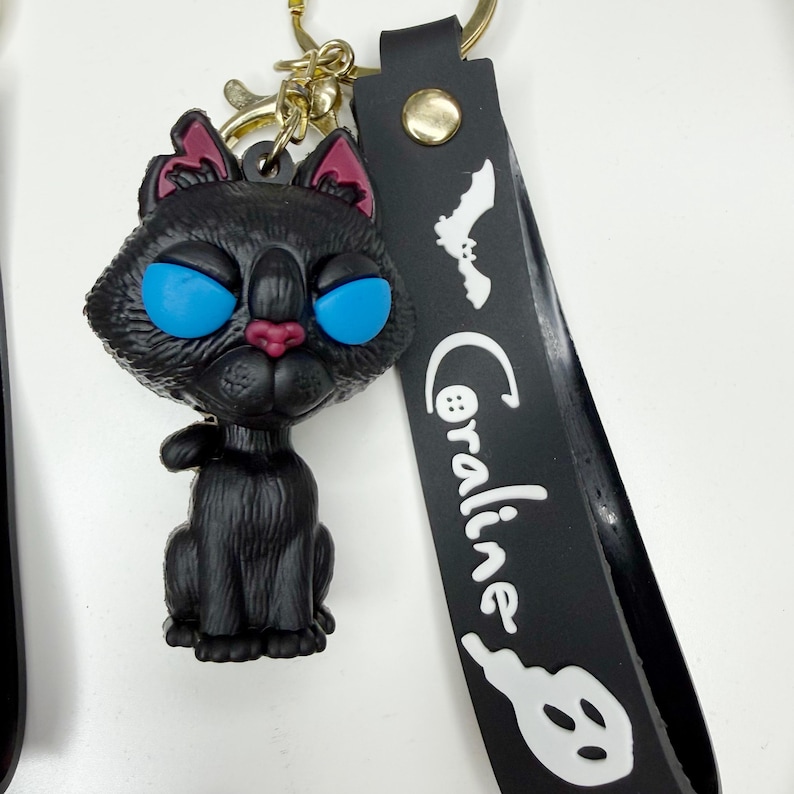 May include: A black cat keyring with blue eyes, pink ears and nose, and a gold-coloured keyring. The black strap has the word "Coraline" in white, along with a bat and a ghost.