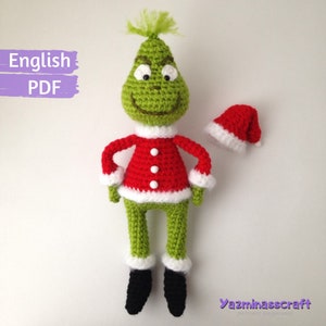 May include: Green crocheted Grinch doll wearing a red and white Santa suit with a white trim. The doll is holding its hands in front of it. The doll is wearing black shoes. The doll is wearing a red Santa hat. The doll is standing on a white background. The text "English PDF" is in the top left corner of the image. The text "Yazminasscraft" is in the bottom right corner of the image.