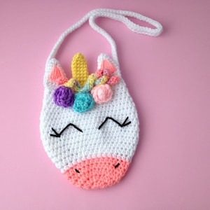 Puede incluir: Bolso de unicornio de crochet blanco con una nariz rosa, flores de hilo de colores arcoíris y una cara bordada en negro.
