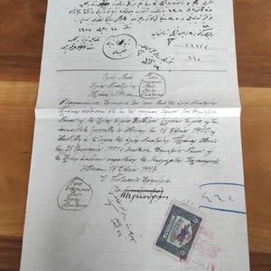 May include: A vintage, handwritten document with text in multiple languages, featuring stamps and signatures. The paper is aged, with visible creases and holes, suggesting historical significance. The document is on a wooden surface.