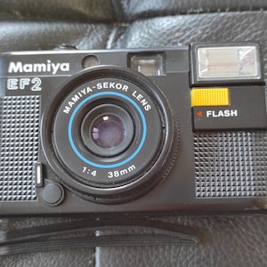 以下が含まれることがあります： 内蔵フラッシュ付きのブラックのMamiya EF2 35mmフィルムカメラ。カメラには、38mmの焦点距離と1:4の絞りを持つMamiya-Sekorレンズが搭載されています。