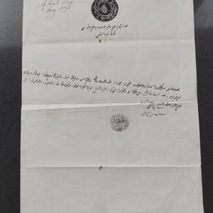 May include: A vintage document with handwritten text and ornate seals. The paper is aged, with visible creases and stains. The text appears to be in a foreign language, with decorative calligraphy and circular seals. The document is folded, suggesting it was once stored.