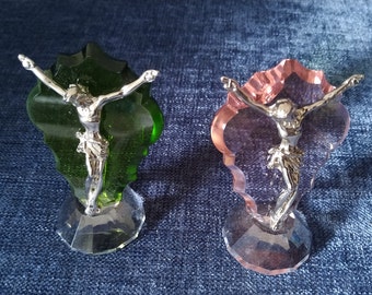 May include: Two crystal figurines of Jesus on the cross. The figurine on the left is on a green crystal base and the figurine on the right is on a pink crystal base.