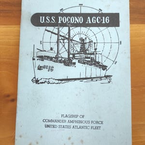 May include: A light blue vintage welcome aboard card for the U.S.S. Pocono AGC-16. The card features a black and white illustration of the ship, a compass design, and text that reads "Flagship of Commander Amphibious Force United States Atlantic Fleet."