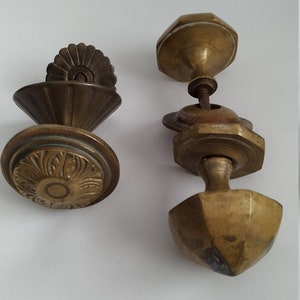 May include: Two antique brass door knobs. One knob has a decorative, ornate design and the other has a simple, geometric design.