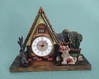 May include: A wooden clock shaped like a house with a green roof and a white clock face. The clock has a brown wooden base and a small dog figurine on the front.