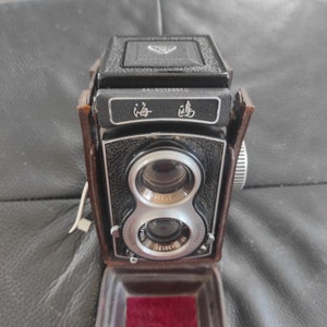 May include: A vintage black and brown twin-lens reflex camera with a leather case. The camera has two lenses, a viewfinder, and a film advance lever. The camera has a label that reads "4A-20160010" and "海鸥".
