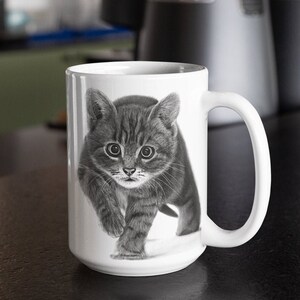 May include: A white ceramic mug with a black and white illustration of a kitten walking towards the viewer. The kitten has a striped pattern and is looking directly at the camera.