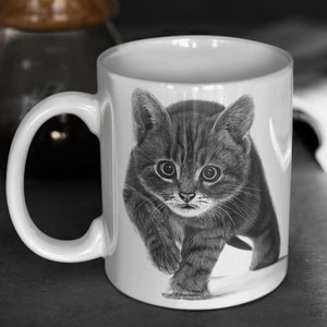 May include: A white ceramic mug with a black and white illustration of a kitten walking towards the viewer. The kitten has a striped pattern and is looking directly at the camera.