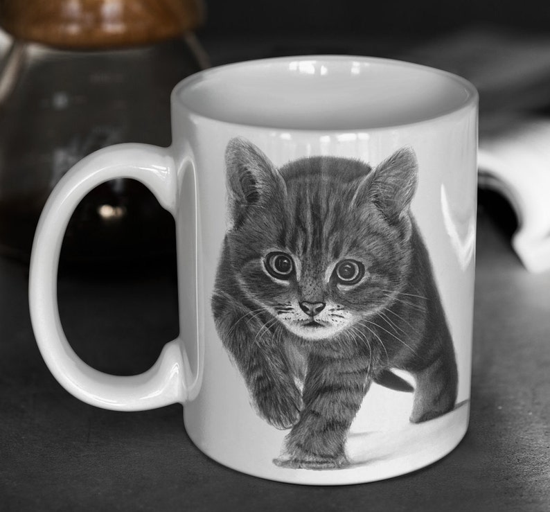 May include: A white ceramic mug with a black and white illustration of a kitten walking towards the viewer. The kitten has a striped pattern and is looking directly at the camera.