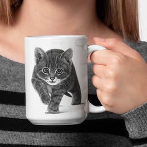 May include: A white ceramic mug with a black and white illustration of a kitten looking directly at the viewer. The kitten is walking towards the viewer with its paws raised.
