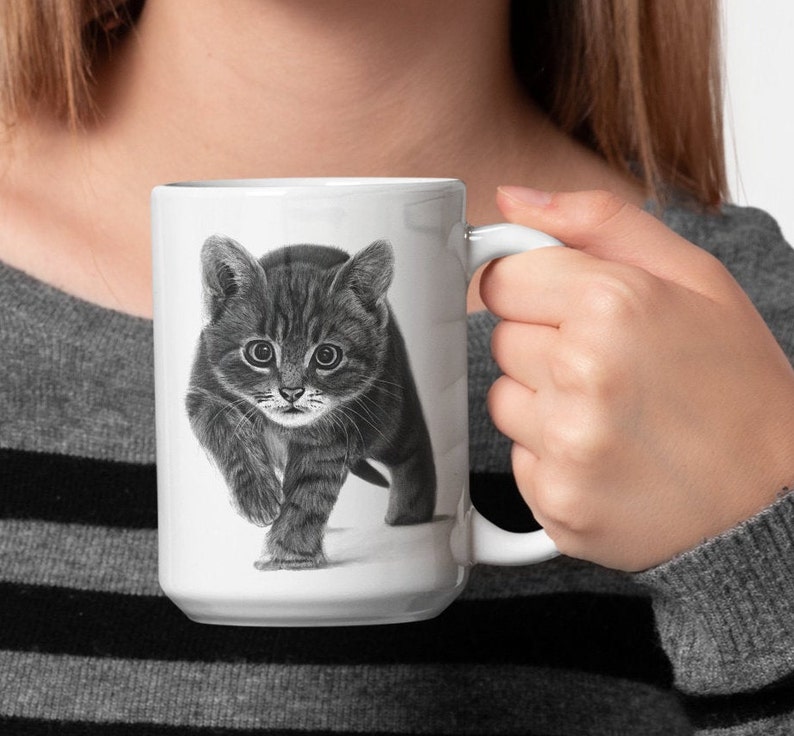 May include: A white ceramic mug with a black and white illustration of a kitten looking directly at the viewer. The kitten is walking towards the viewer with its paws raised.