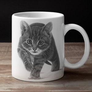 May include: A white ceramic mug with a black and white drawing of a tabby kitten looking directly at the viewer. The kitten is walking towards the viewer with its paws raised.