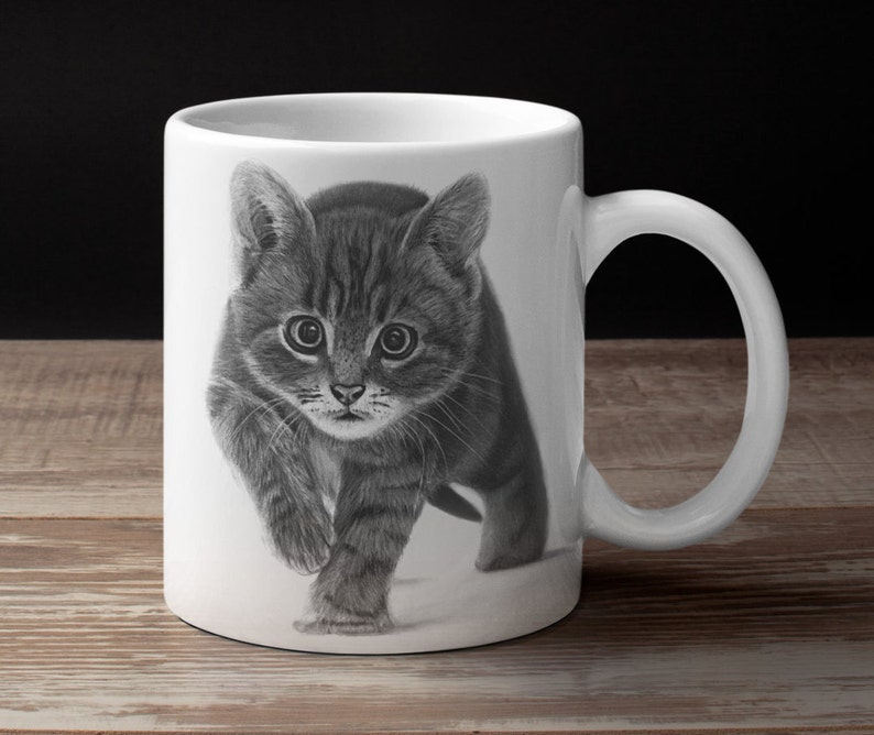 May include: A white ceramic mug with a black and white drawing of a tabby kitten looking directly at the viewer. The kitten is walking towards the viewer with its paws raised.