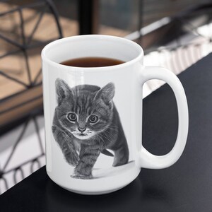 May include: A white ceramic mug with a black and white illustration of a kitten walking towards the viewer. The mug has a black handle and is filled with dark brown coffee.