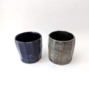 Peut inclure: Deux tasses en céramique faites à la main. L'une est bleu marine foncé, l'autre est un mélange de gris et de marron. Les deux ont un design à facettes et un intérieur sombre. Les tasses sont probablement pour boire des boissons.