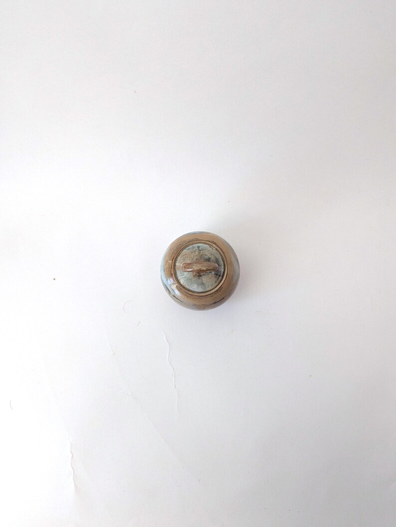 Puede incluir: Un peque&ntilde;o pomo redondo de estilo antiguo con acabado met&aacute;lico. El pomo tiene una zona central ligeramente empotrada con un color m&aacute;s claro, rodeada de metal m&aacute;s oscuro y envejecido. El pomo es probablemente para un armario o caj&oacute;n.