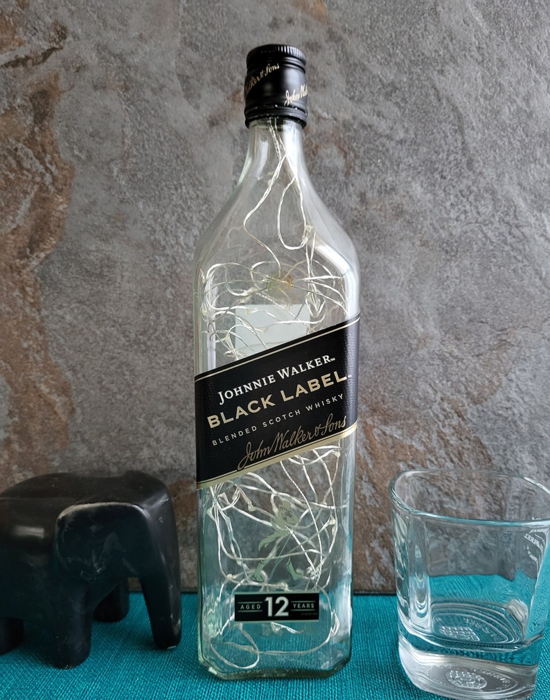 Puede incluir: Una botella de vidrio transparente con una etiqueta negra que dice "JOHNNIE WALKER. BLACK LABEL BLENDED SCOTCH WHISKY John Walker & Sons AGED 12 YEARS". La botella est&aacute; llena de luces blancas.