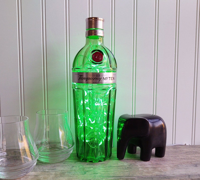 May include: A green glass bottle with a label that reads "Tanqueray No. Ten" and is filled with green fairy lights. The bottle is sitting on a white surface with a black elephant figurine in front of it.