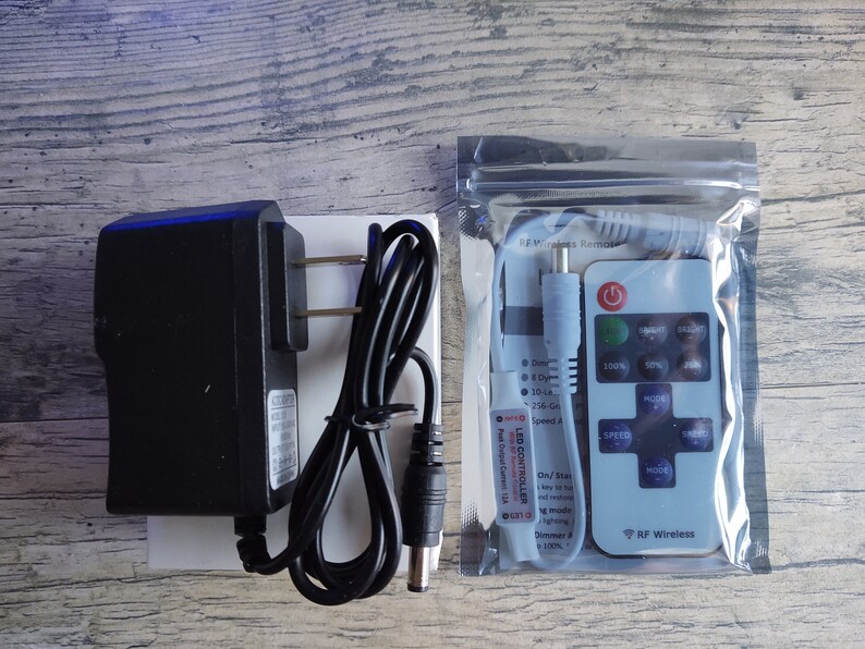 May include: A black power adapter with a cord and a white plug, and a white remote control with a black button and a black power cord. The remote control has the text "RF Wireless" and "LED CONTROLLER" printed on it.
