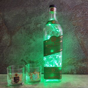 May include: A clear glass bottle with a green label that says "Johnnie Walker Pure Malt Scotch Whisky" and "15" is filled with green fairy lights. Two clear glass tumblers with gold accents are in front of the bottle.