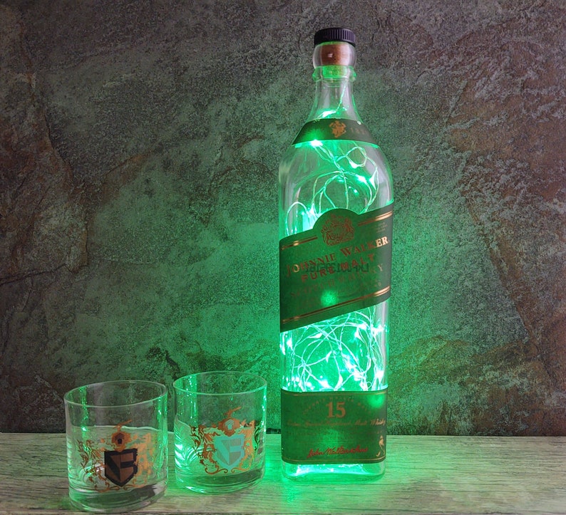 May include: A clear glass bottle with a green label that says "Johnnie Walker Pure Malt Scotch Whisky" and "15" is filled with green fairy lights. Two clear glass tumblers with gold accents are in front of the bottle.