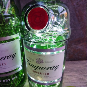 May include: A green glass bottle with a red label and white lights wrapped around it. The label says "Tanqueray Imported" and "Distilled Four Times from the World's Finest Botanicals".