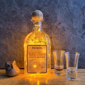 May include: A clear glass bottle with a cork top and string lights inside. The bottle is labeled "PATRÓN. ESTATE RELEASE" and is sitting on a wooden surface with two shot glasses.