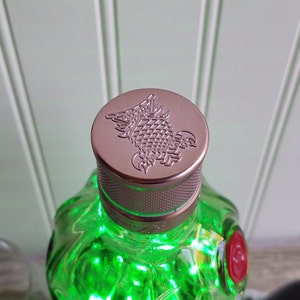 May include: A green glass bottle with a silver and copper colored cap. The cap has a detailed embossed design of a mythical creature.