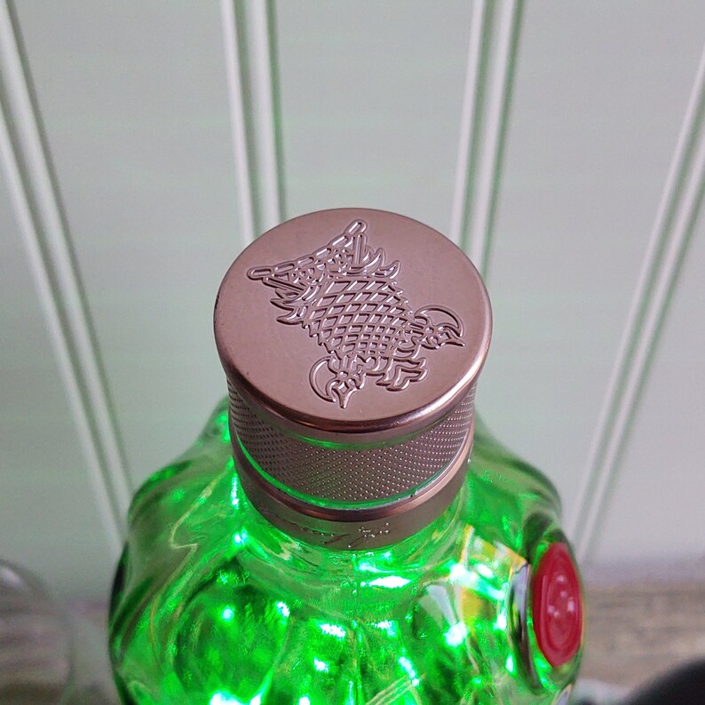 May include: A green glass bottle with a silver and copper colored cap. The cap has a detailed embossed design of a mythical creature.