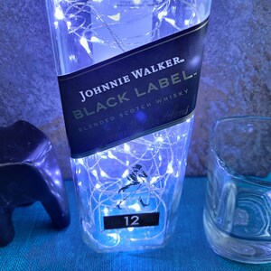 Puede incluir: Una botella de vidrio transparente con una etiqueta negra que dice "JOHNNIE WALKER BLACK LABEL BLENDED SCOTCH WHISKY" envuelta en luces blancas de hadas. La botella est&aacute; iluminada con luz azul. El n&uacute;mero "12" es visible en la etiqueta.