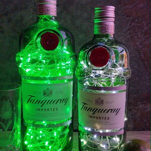 May include: Two empty glass bottles with green and white string lights inside. The bottles are labeled "Tanqueray Imported" and have a red seal. The bottles are sitting on a wooden surface.