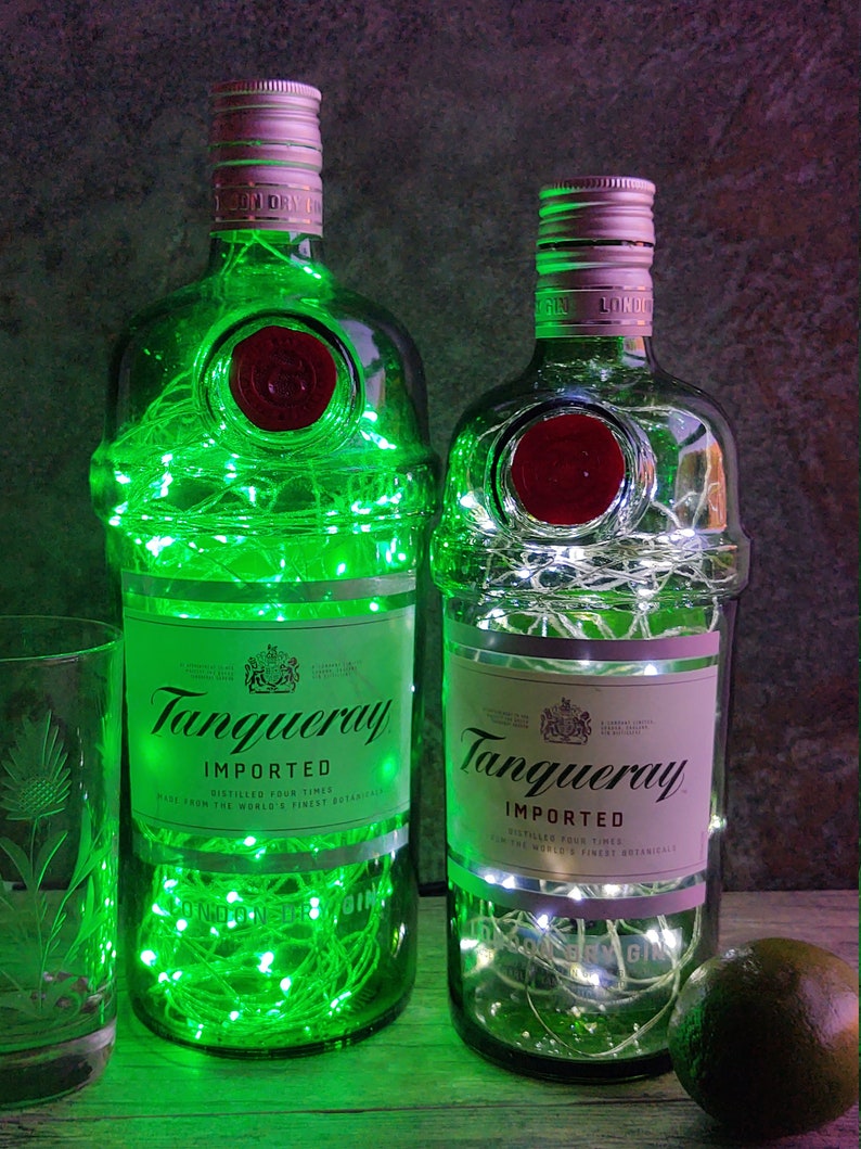 May include: Two empty glass bottles with green and white string lights inside. The bottles are labeled "Tanqueray Imported" and have a red seal. The bottles are sitting on a wooden surface.