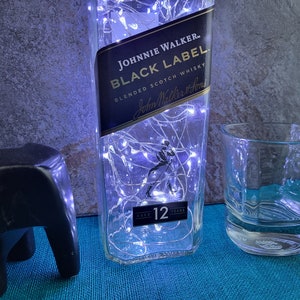 Puede incluir: Una botella de vidrio transparente con una etiqueta negra que dice "JOHNNIE WALKER. BLACK LABEL BLENDED SCOTCH WHISKY John Walker & Sons AGED 12 YEARS". La botella est&aacute; decorada con luces blancas. Un vaso de vidrio transparente se encuentra al lado de la botella.