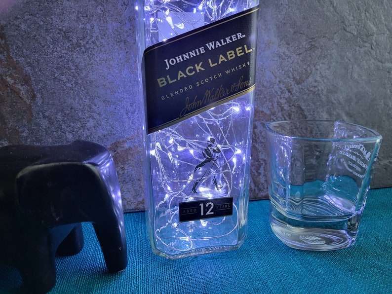 Puede incluir: Una botella de vidrio transparente con una etiqueta negra que dice "JOHNNIE WALKER. BLACK LABEL BLENDED SCOTCH WHISKY John Walker & Sons AGED 12 YEARS". La botella est&aacute; decorada con luces blancas. Un vaso de vidrio transparente se encuentra al lado de la botella.