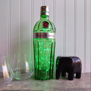 May include: A green glass bottle of Tanqueray No. Ten gin with a string of green fairy lights wrapped around it. The bottle is sitting on a wooden surface next to two clear glass tumblers and a black elephant figurine.
