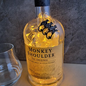 May include: A clear glass bottle of Monkey Shoulder blended malt scotch whisky with string lights inside. The bottle has a black monkey emblem and a label with text. A clear glass is in the foreground.