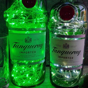 May include: Two green glass bottles with white string lights inside. The bottles are labeled "Tanqueray Imported" and have a red seal on the top. The bottles are sitting on a wooden surface.
