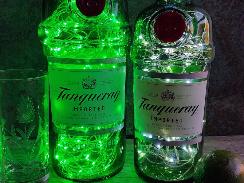 May include: Two green glass bottles with white string lights inside. The bottles are labeled "Tanqueray Imported" and have a red seal on the top. The bottles are sitting on a wooden surface.
