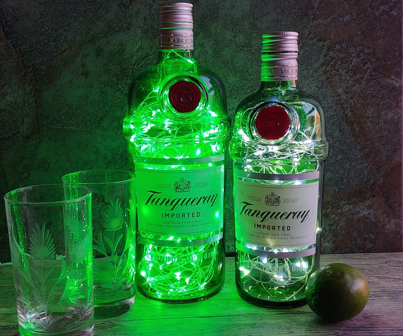 May include: Two clear glass bottles of Tanqueray gin, decorated with green and white fairy lights, sitting on a wooden surface with two clear glasses and a lime.