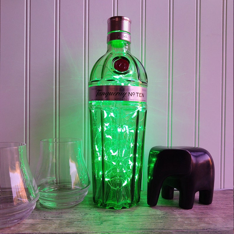 May include: A green glass bottle of Tanqueray No Ten gin with a silver label and a black elephant figurine. The bottle is illuminated with green fairy lights.