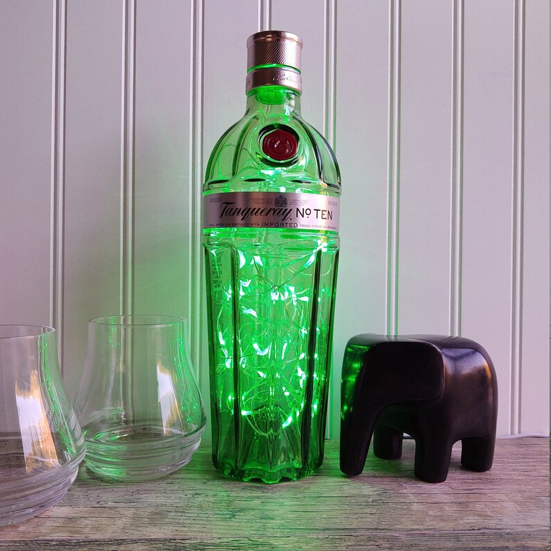 May include: A green glass bottle of Tanqueray No. Ten gin with a string of white lights wrapped around it. The bottle is sitting on a wooden surface with two clear glass tumblers and a black elephant figurine.