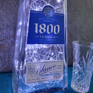 May include: A clear glass bottle with a blue label that reads "1800 Tequila" and "100% Agave Azul". The bottle is decorated with white string lights. Two clear shot glasses are in the foreground.