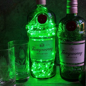 May include: Two green glass bottles of Tanqueray London Dry Gin with green fairy lights inside. The bottles are on a wooden surface with a clear glass with a thistle design.