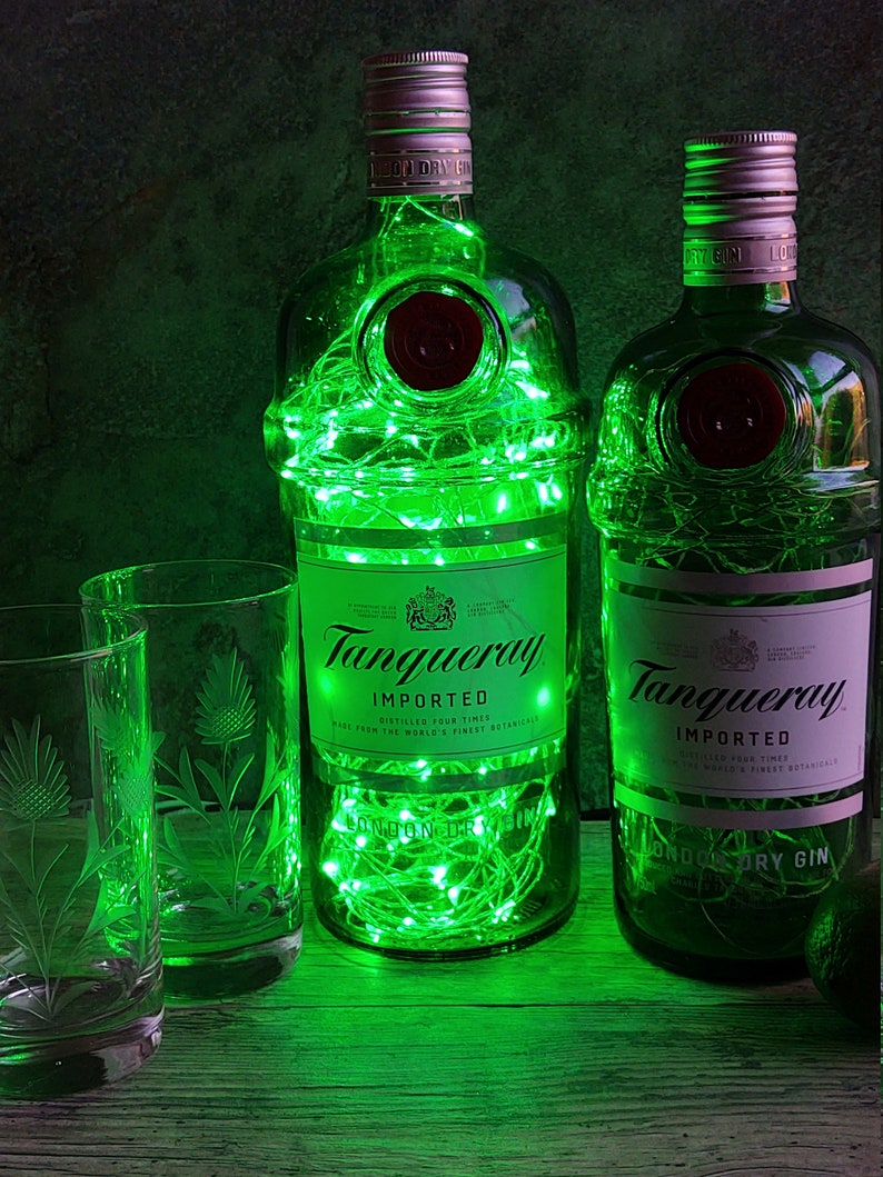 May include: Two green glass bottles of Tanqueray London Dry Gin with green fairy lights inside. The bottles are on a wooden surface with a clear glass with a thistle design.