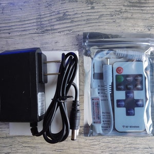 Puede incluir: Un adaptador de corriente negro con un cable y un enchufe blanco, y un mando a distancia blanco con un bot&oacute;n negro y un cable de alimentaci&oacute;n negro. El mando a distancia tiene el texto "RF Wireless" y "LED CONTROLLER" impreso en &eacute;l.