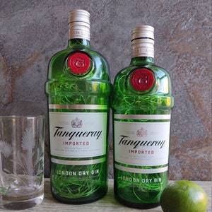 May include: Two green glass bottles of Tanqueray London Dry Gin, one larger than the other, with the brand name and text "IMPORTED" and "DISTILLED FOUR TIMES" printed on the labels. The bottles are illuminated with white string lights.