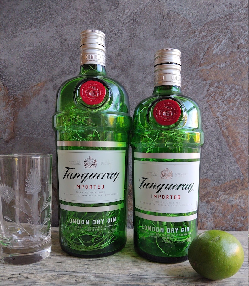 May include: Two green glass bottles of Tanqueray London Dry Gin, one larger than the other, with the brand name and text "IMPORTED" and "DISTILLED FOUR TIMES" printed on the labels. The bottles are illuminated with white string lights.